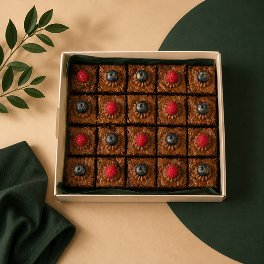 A top-down view of a square box of chocolate brownies, each topped with a piped flower-like swirl of frosting and a red raspberry or blueberry in the center. The box is placed on a beige surface with a dark green circular shape, a folded dark green napkin, and a leafy branch in the corners.